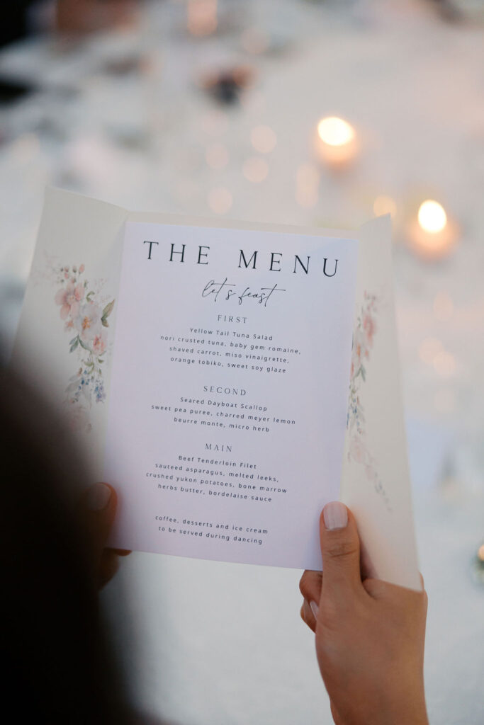 Person holding a menu with floral design, listing courses such as yellowtail tuna salad, seared scallop, and kingfish with dessert options mentioned.