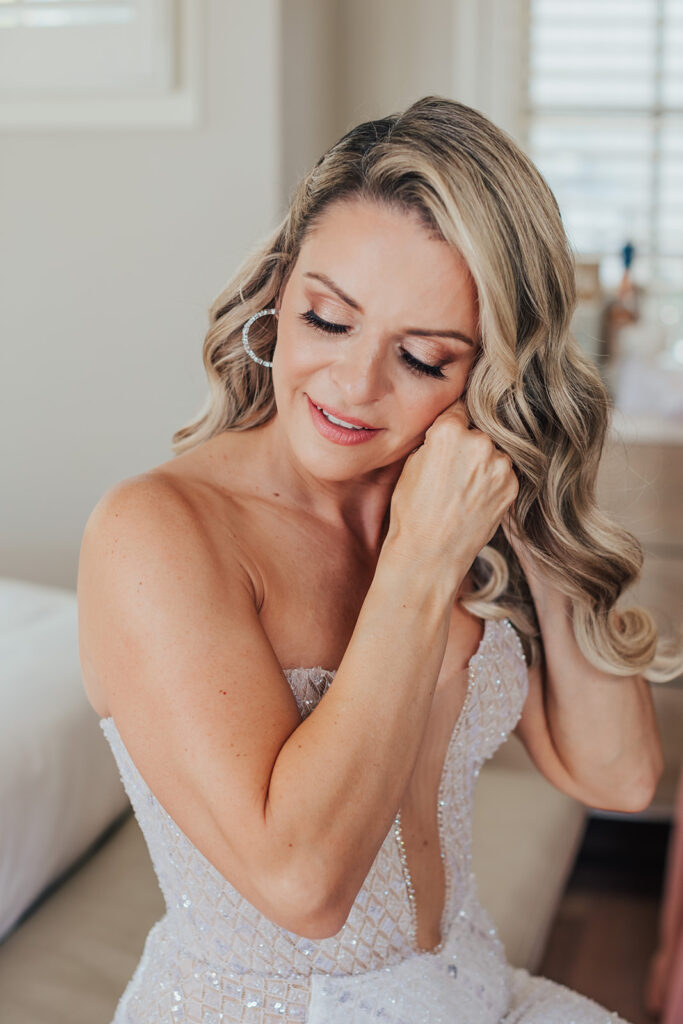 A woman with curled blonde hair adjusts her earring while wearing a strapless, beaded gown.