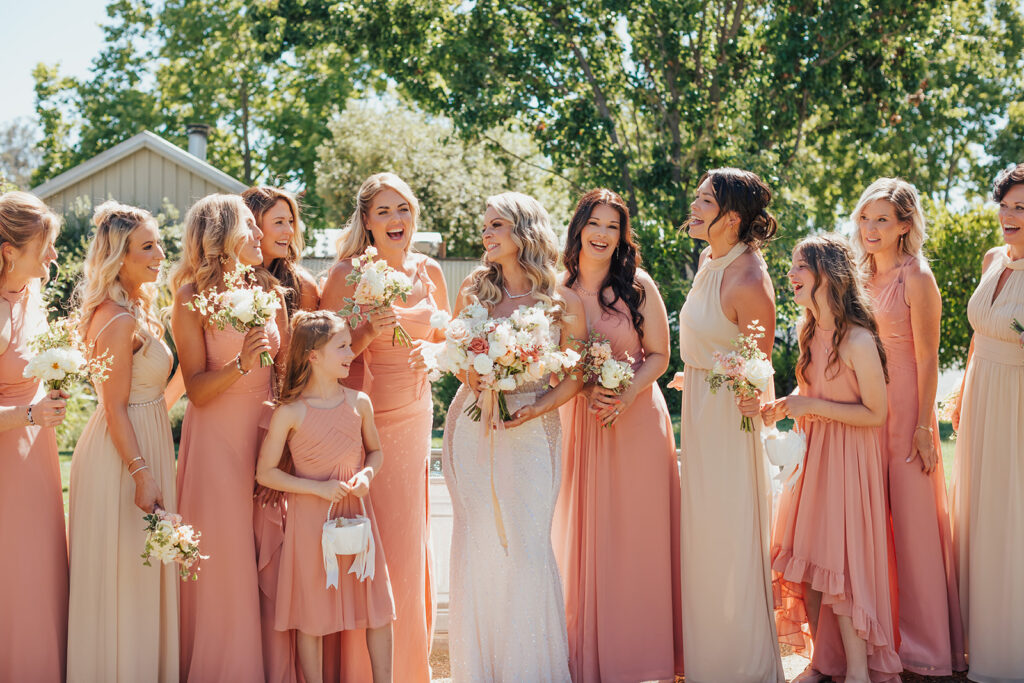 A bride stands with her bridesmaids, all dressed in peach and beige dresses, holding floral bouquets, outdoors on a sunny day.