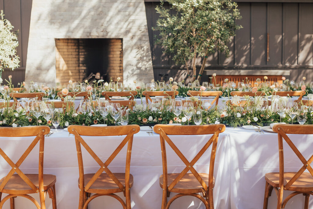 Andria and James' wedding featured an elegant outdoor dining setup with long tables draped in white cloths, adorned with floral centerpieces. Wooden chairs surrounded the scene, while a fireplace flickered warmly in the background, creating a perfect ambience.