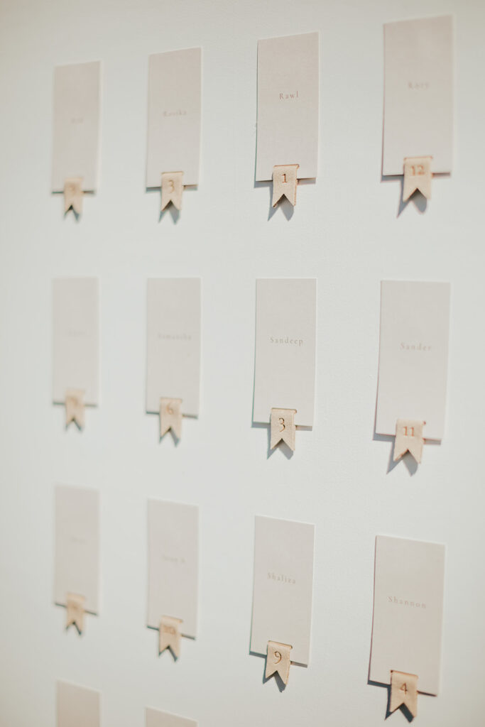 A wall display for Andria and James' wedding features 15 name cards with numbers, each attached to a small wooden clip. Names and numbers elegantly adorn light-colored cards arranged in a charming grid pattern.