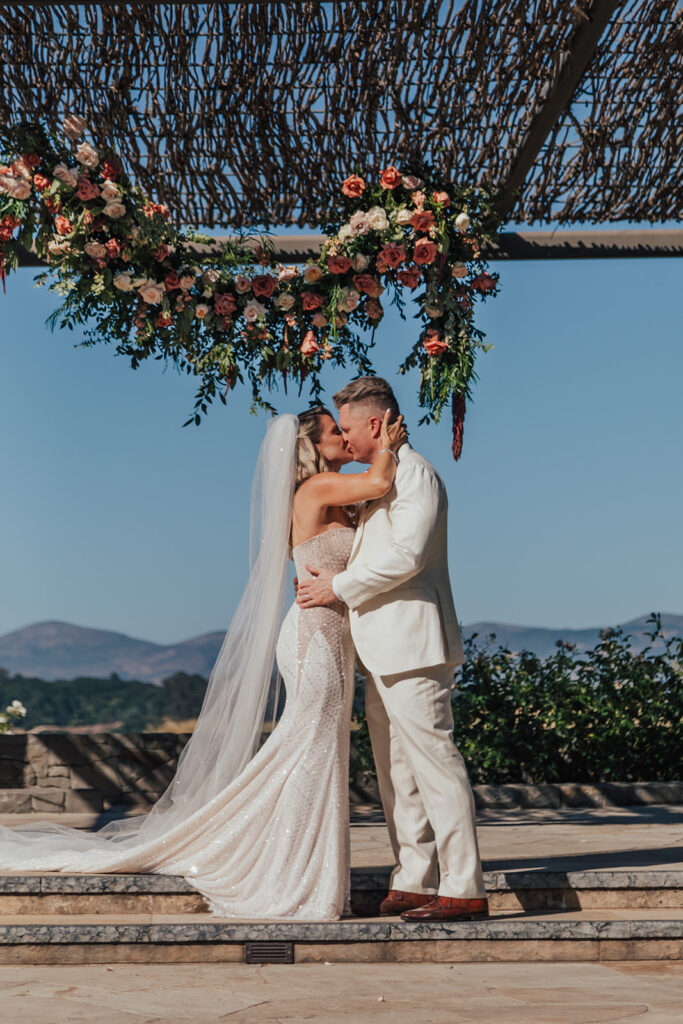 A couple in wedding attire kisses under a floral arch with a mountainous backdrop.