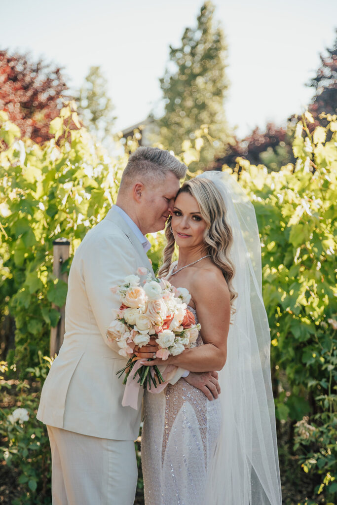 Bride and groom embrace in a vineyard, with the groom in a white suit and the bride holding a bouquet of flowers.