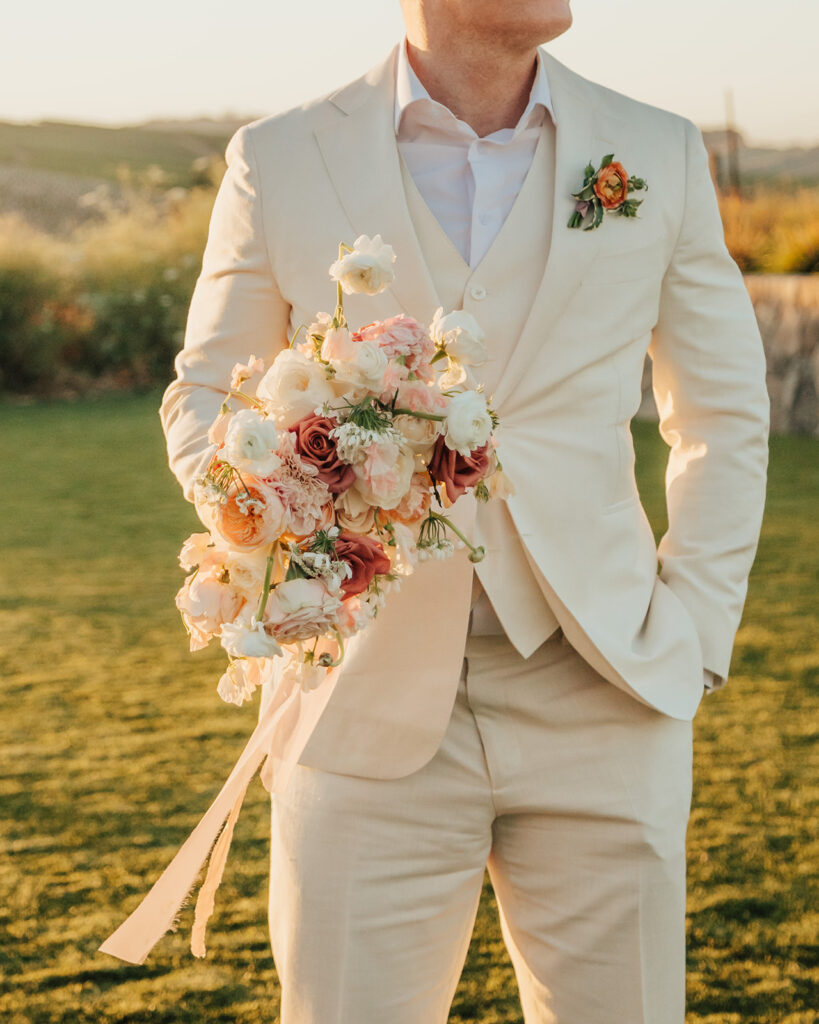 A person in a light-colored suit holds a floral bouquet, standing on grass with a blurred landscape in the background.