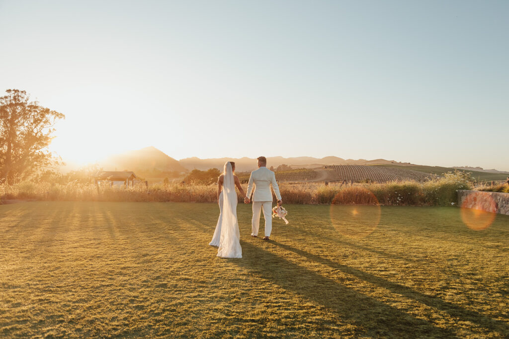 A couple in wedding attire walks hand in hand across a grassy field at sunset.