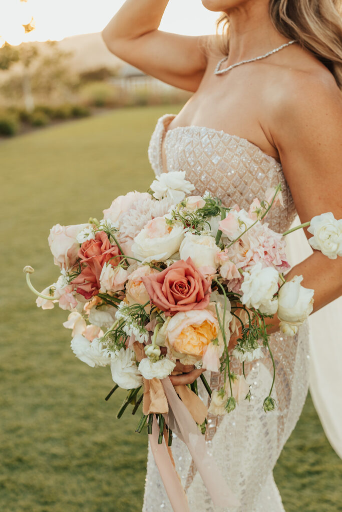 A bride holds a bouquet of pink, peach, and white flowers. She wears a strapless beaded gown and a necklace. The background features green grass and trees.