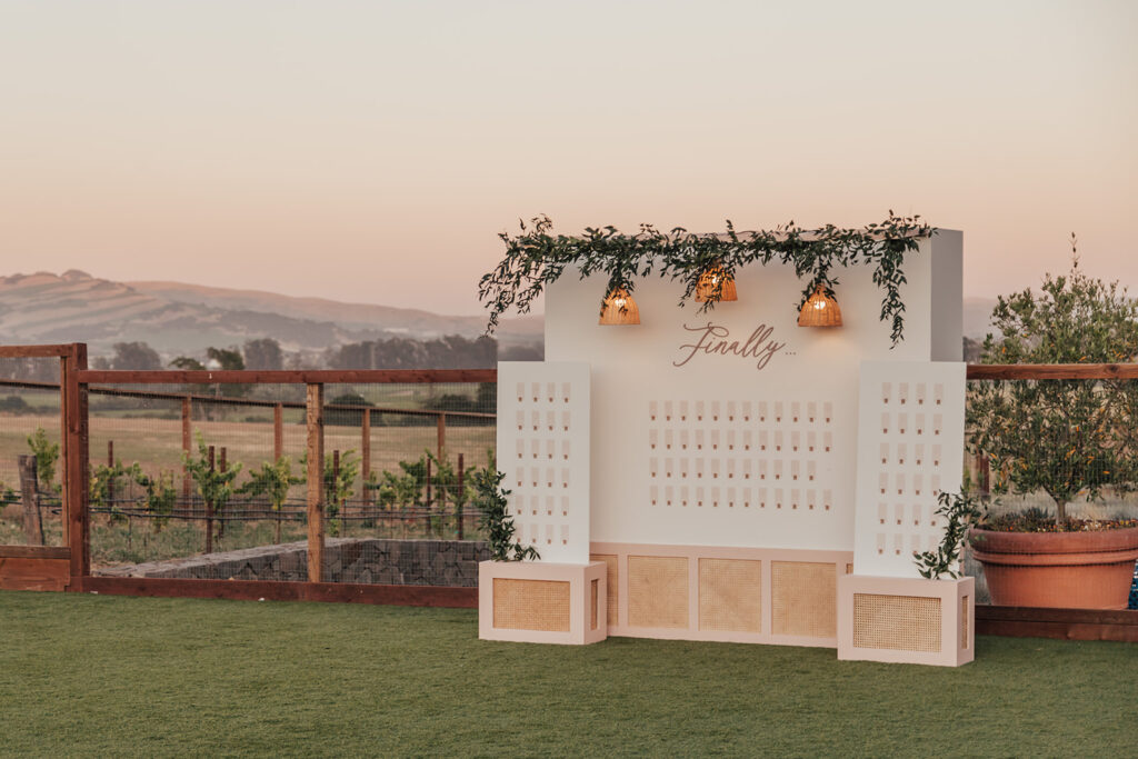 Outdoor seating chart display with greenery and hanging lights, labeled "Finally," set against a countryside backdrop at sunset, for Andria and James' wedding.