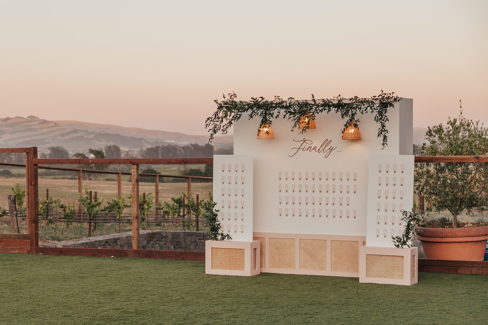 Outdoor seating chart display with greenery and hanging lights, labeled "Finally," set against a countryside backdrop at sunset, for Andria and James' wedding.