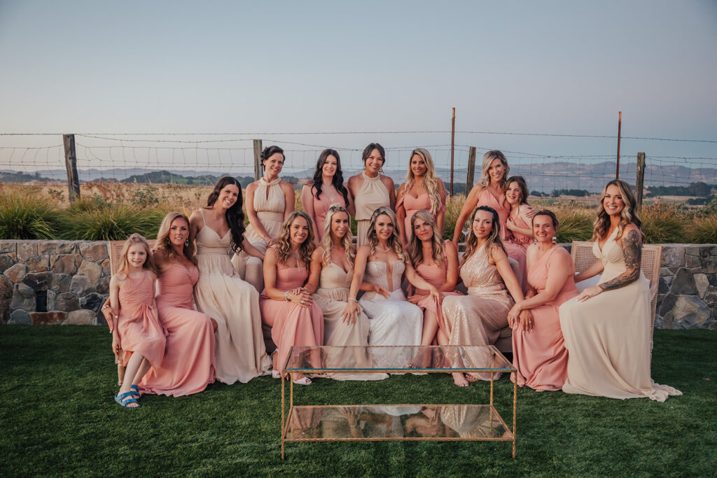 A group of women and a girl in pink and beige dresses sit and stand together outdoors. A stone wall and fence are in the background.