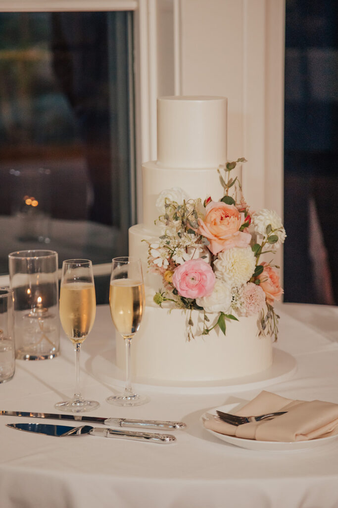 A three-tier white cake adorned with pink and white flowers next to two champagne flutes, candles, cutlery, and a folded napkin on a table.