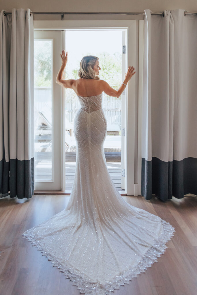 A woman in a sequin-embellished, strapless gown stands facing a glass door with curtains, her back to the camera.