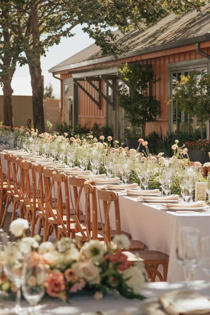 A long outdoor dining table is set with white tablecloths, wooden chairs, glassware, and floral centerpieces, next to a rustic building surrounded by greenery.