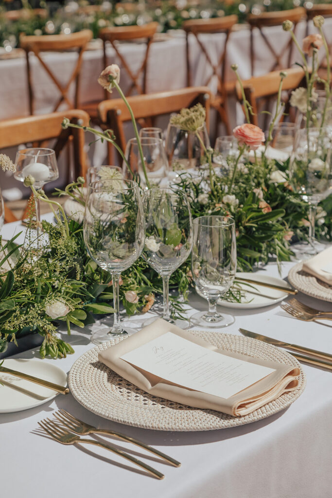 A table set for a formal outdoor event with white tablecloth, greenery centerpieces, wine glasses, plates, and silverware.