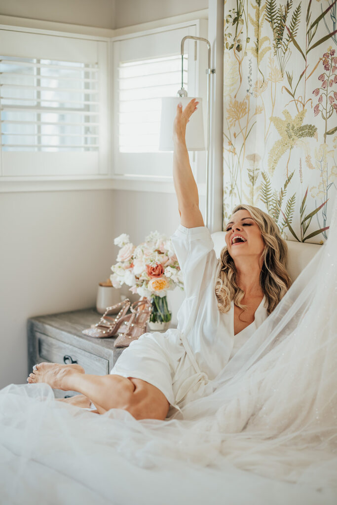 A woman in a white robe sits joyfully on a bed, reaching for a lamp. A bouquet and shoes are on the bedside table.
