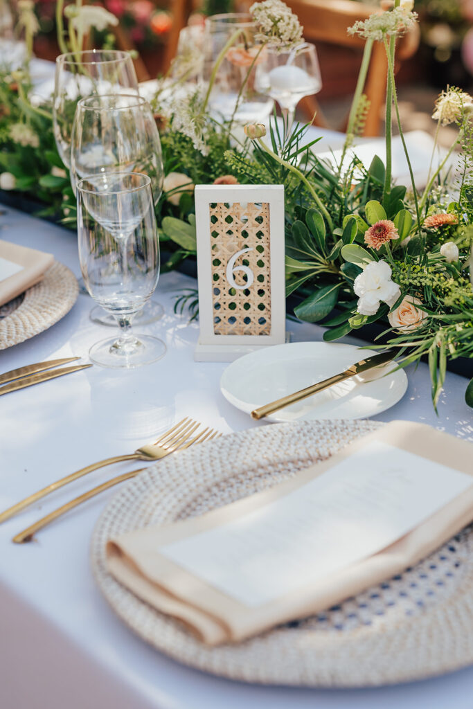 Elegant table setting at Andria and James' wedding with gold cutlery, white plates on woven placemats, wine glasses, and a floral centerpiece. A white card with the number 6 is displayed.