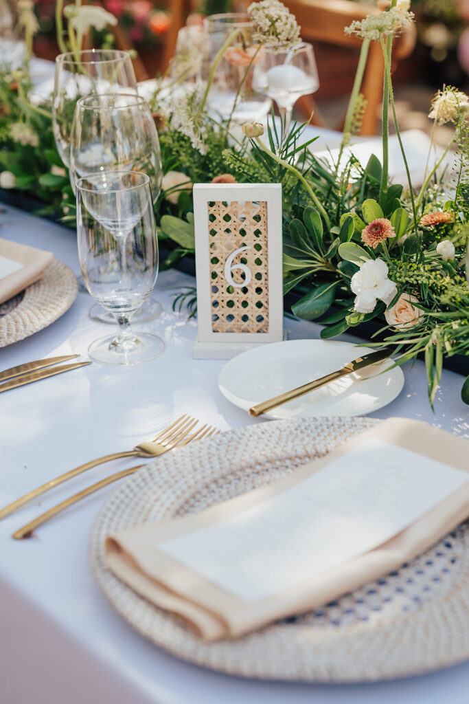 Elegant table setting with gold utensils, woven placemats, and white plates. A floral centerpiece runs along the middle, with a table number card displayed prominently.