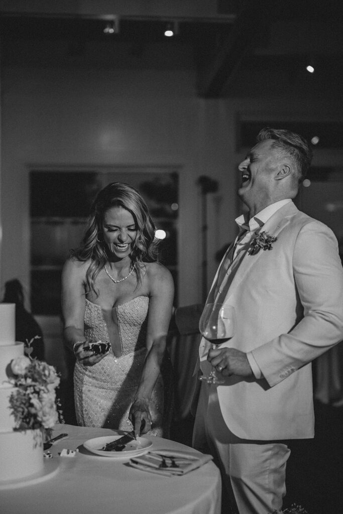 A couple laughs while cutting a cake at a wedding reception; the woman holds a piece of cake, and the man holds a glass.