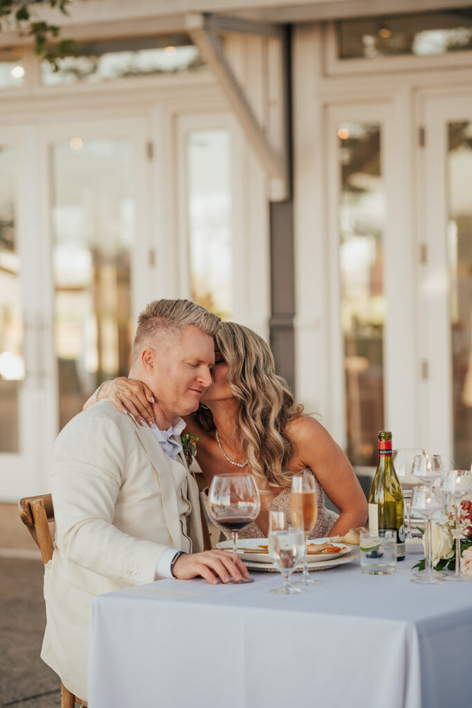 A couple sits at a table, embracing with closed eyes. A bottle of wine, wine glasses, and plates are on the table. The background features a building with large windows and doors.