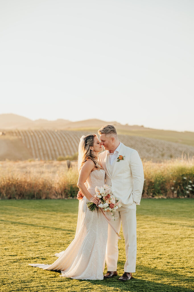 A couple dressed in wedding attire stands closely on a sunlit grassy field, with vineyards in the background.