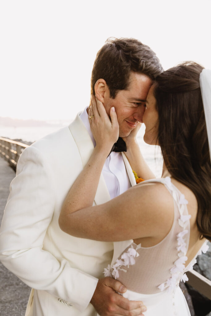 A bride and groom in formal attire embrace closely, touching foreheads and smiling, on a boardwalk.