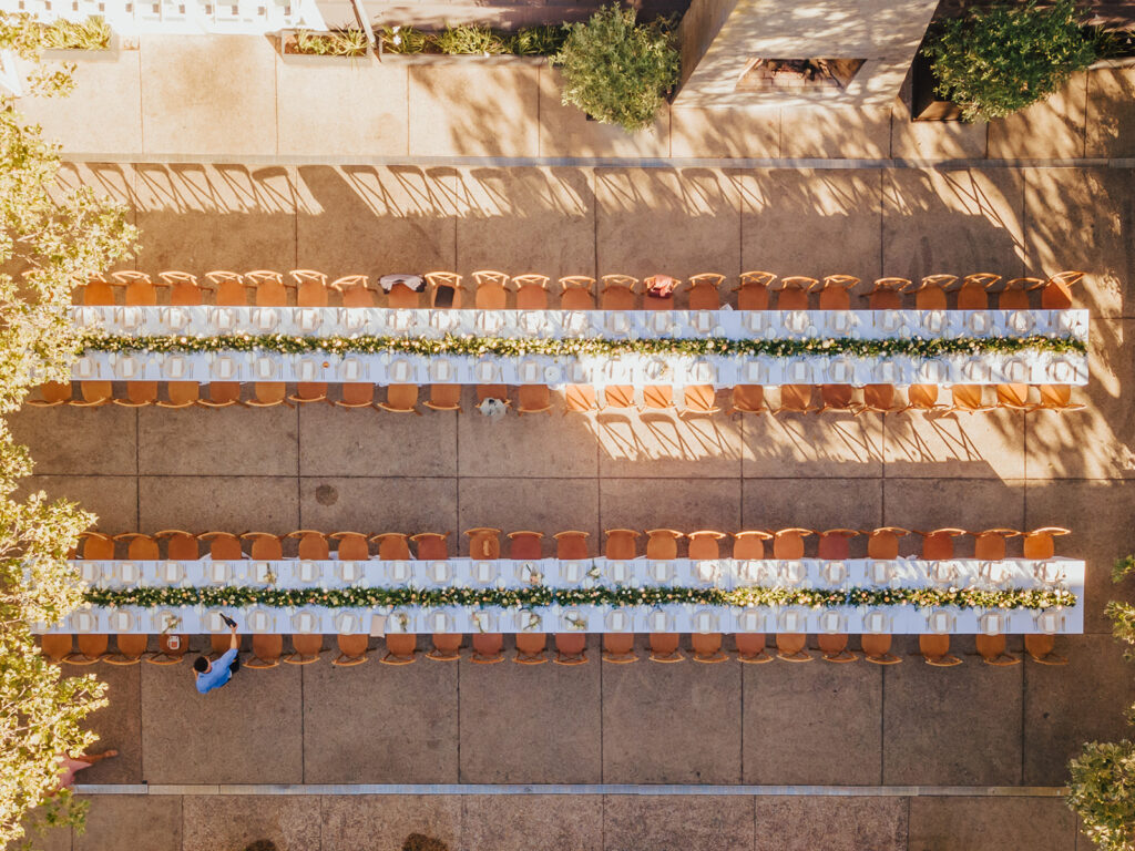 Aerial view of two long tables set up outdoors with white tablecloths and floral centerpieces, surrounded by wooden chairs.
