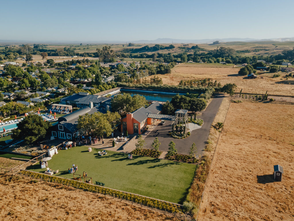 Aerial view of a rural property with buildings, a lawn area, and surrounding fields. People are visible on the lawn, and hills extend into the distance.