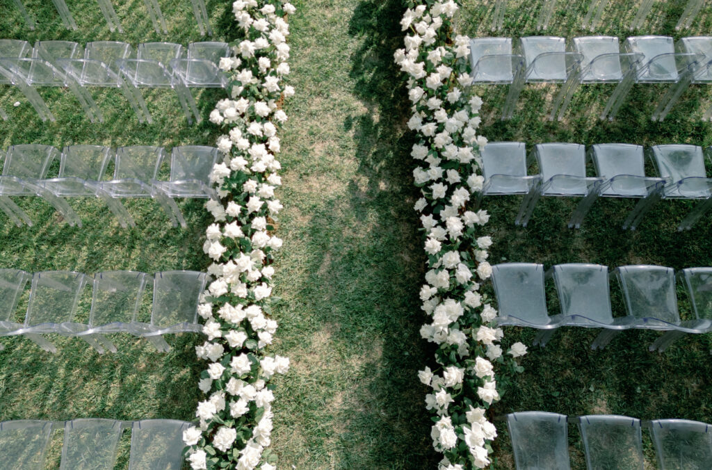 Rows of transparent chairs are arranged for an outdoor event. White flowers line the grassy aisle in the center.