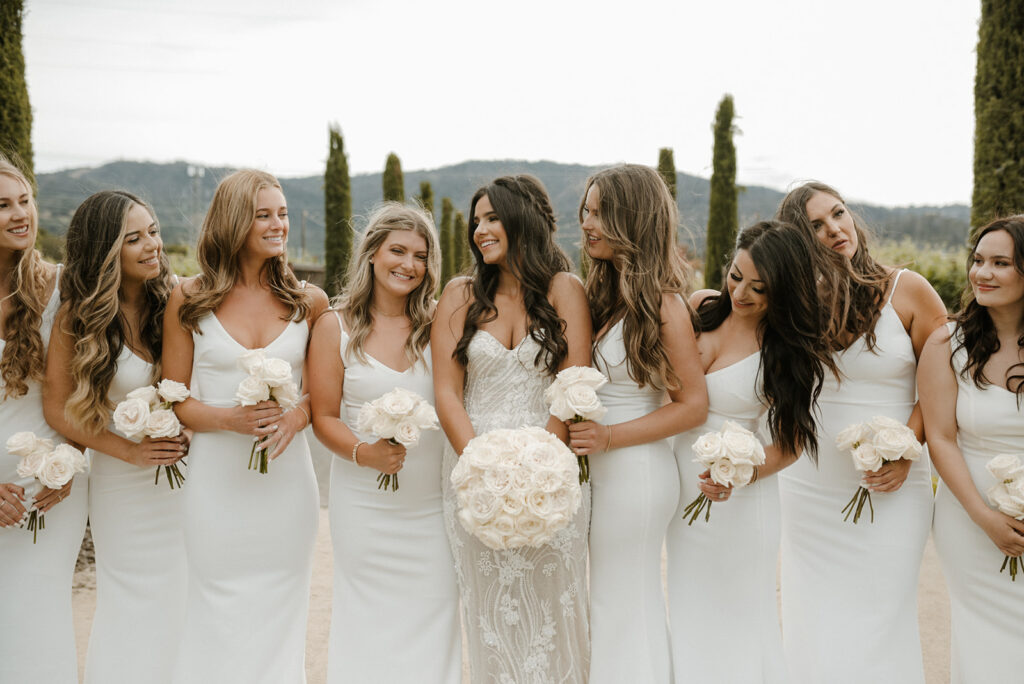 A bride in a white dress stands with eight bridesmaids, all wearing white dresses and holding white rose bouquets. They are outdoors with tall trees in the background.