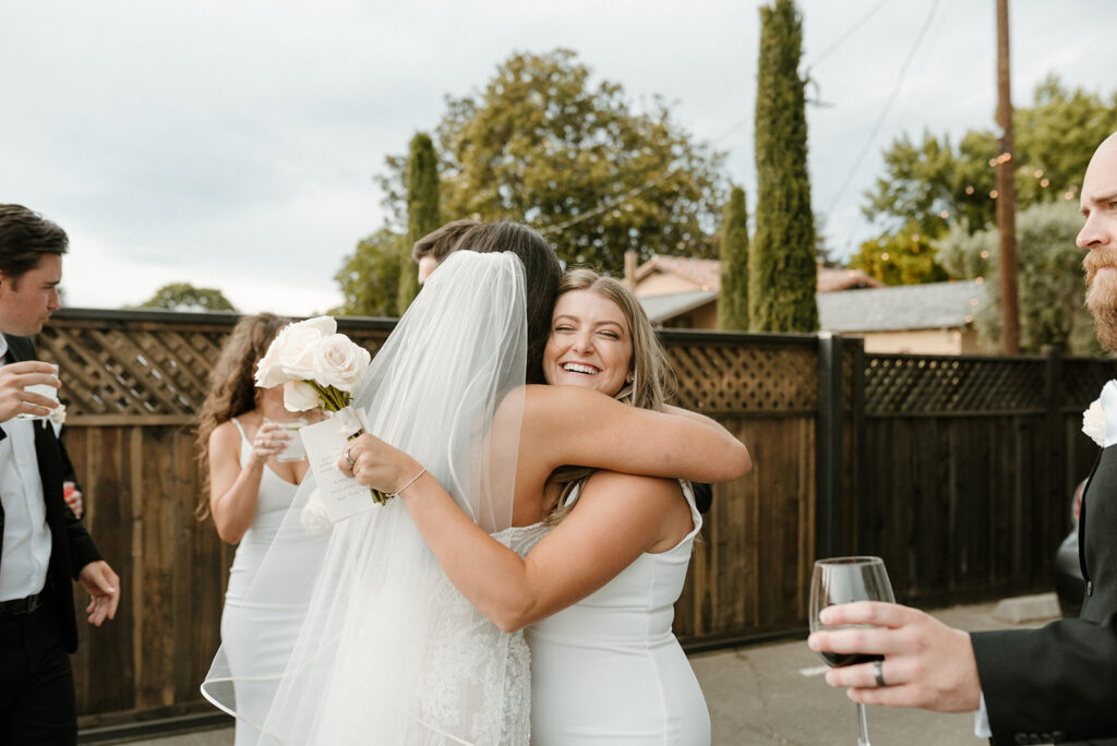 A bride in a white dress and veil hugs a bridesmaid holding a bouquet, while other people in formal attire hold drinks nearby.