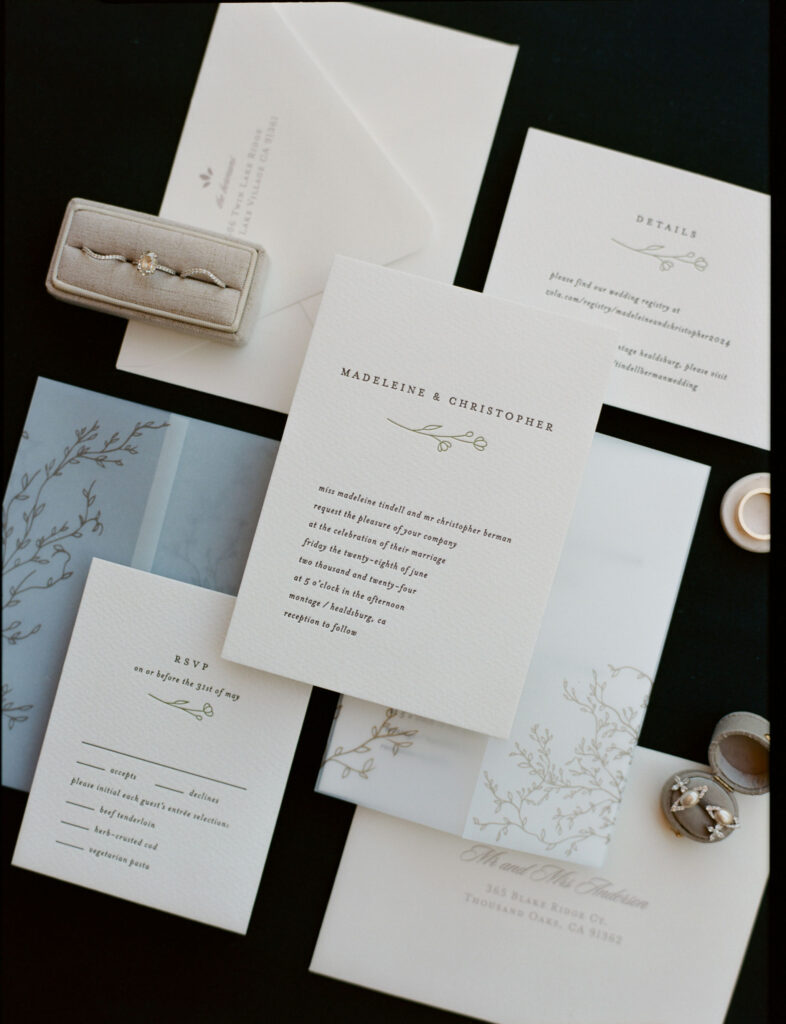 A set of elegant wedding invitations and RSVP cards arranged on a dark surface, accompanied by a ring box and decorative envelopes.