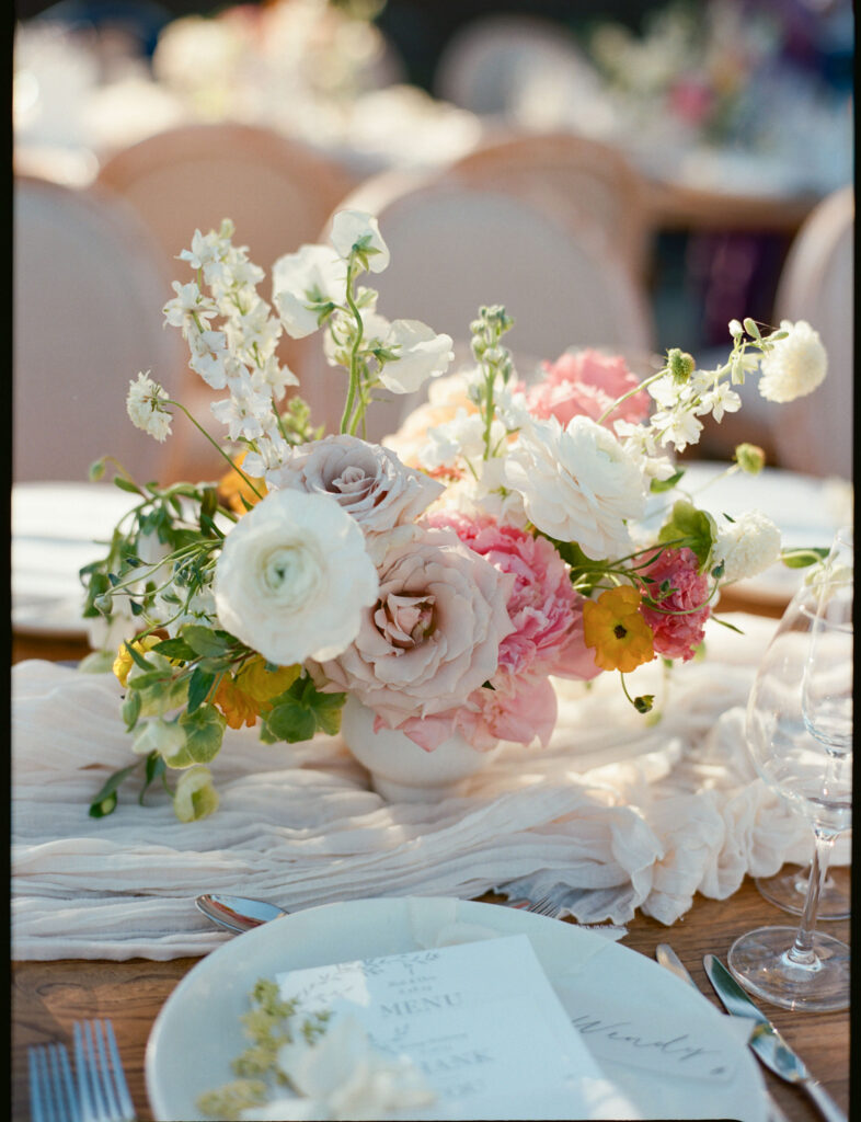 A floral centerpiece featuring pink, white, and yellow flowers in a vase on a table set with plates and menus.