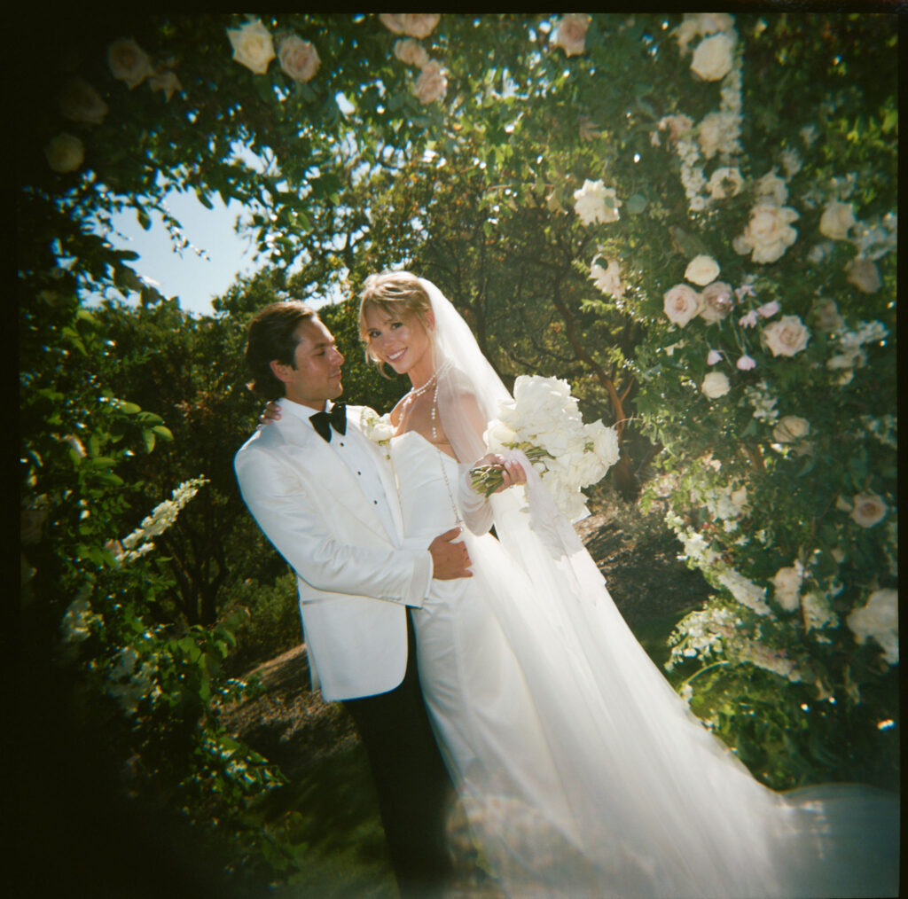 Bride and groom in wedding attire stand under a floral arch, smiling at each other, surrounded by greenery and white flowers.
