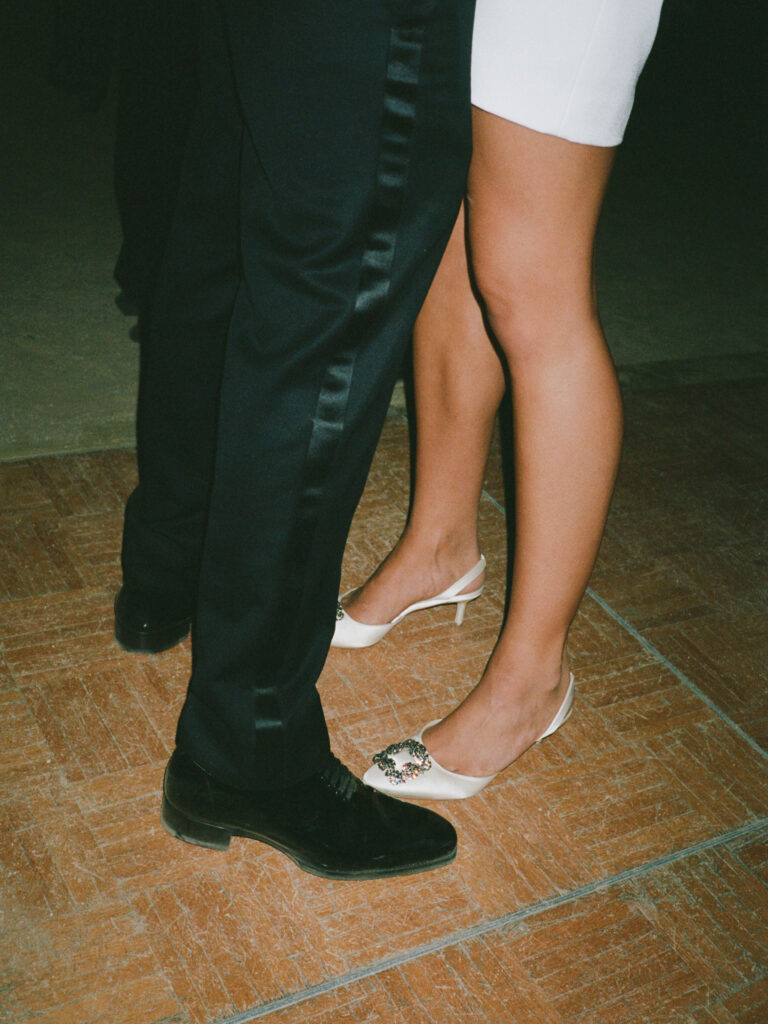 A couple dances closely on a tiled floor, with the man in dark pants and the woman in a white dress and white heels.