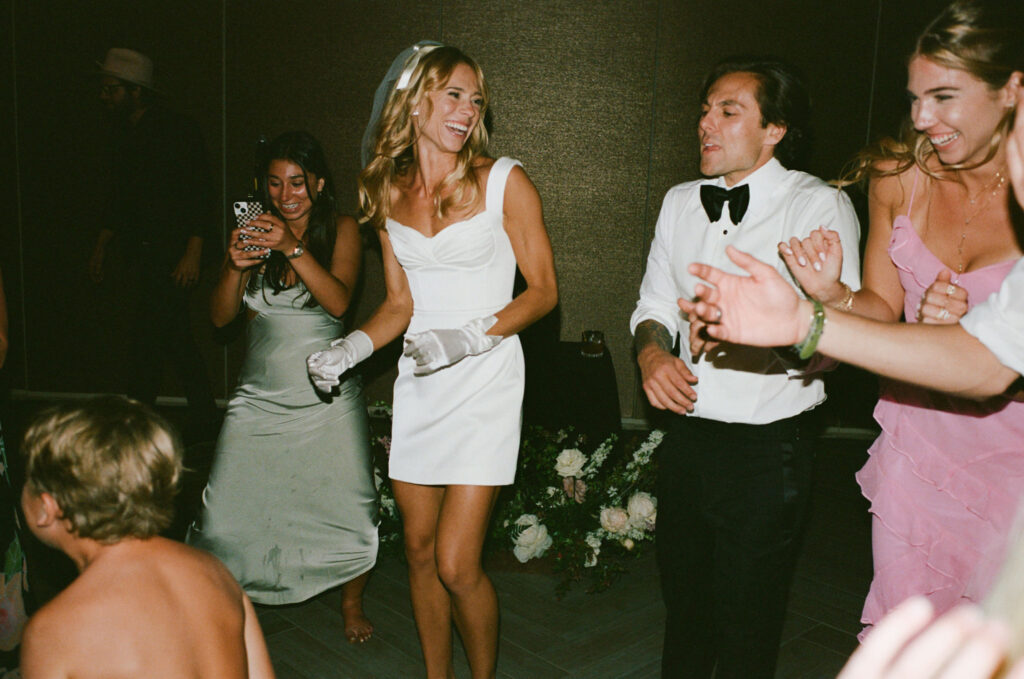 A group of people in formal attire dancing at an indoor event. A woman in a white dress and gloves is smiling in the center.