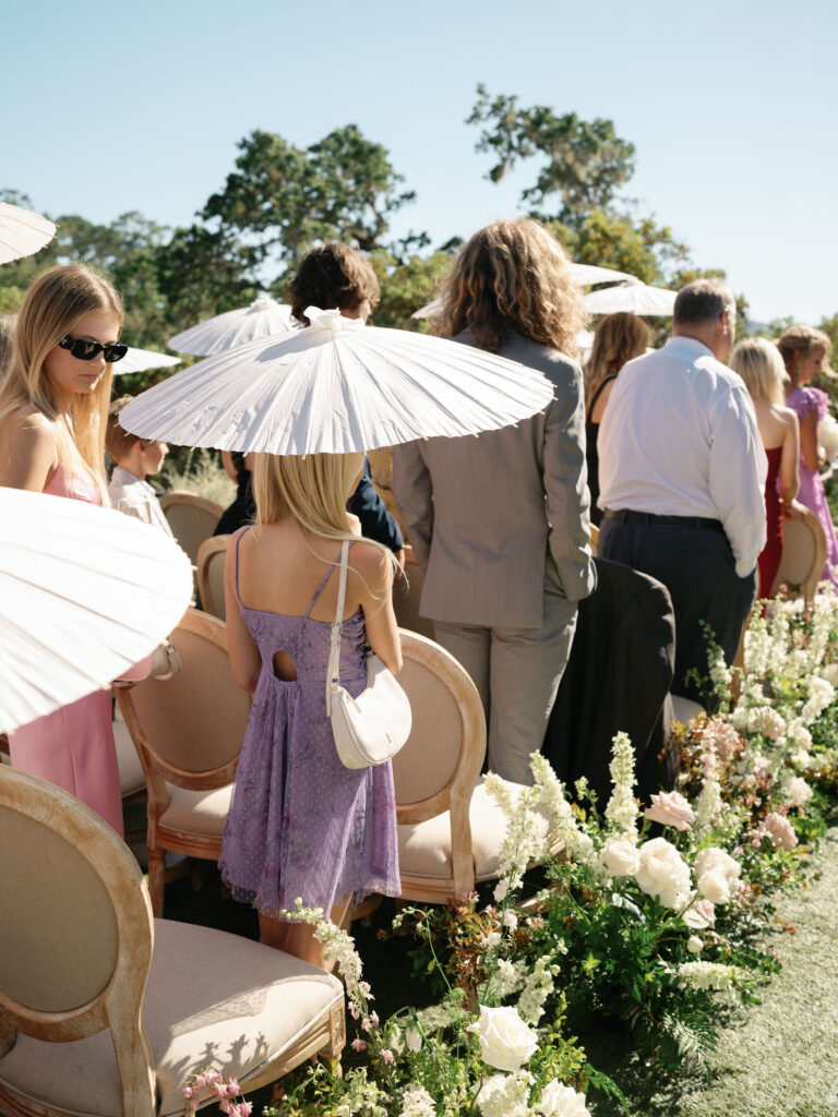 Wedding ceremony guests stand outdoors with parasols, surrounded by floral arrangements.