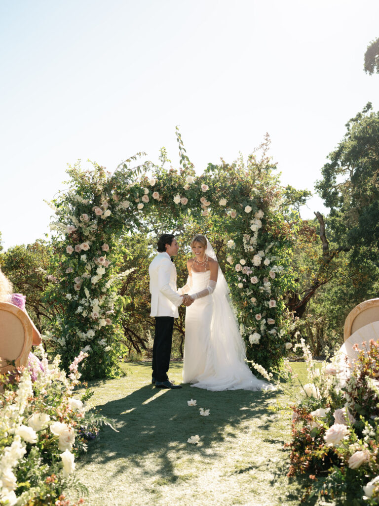 A couple stands under a floral arch during an outdoor wedding ceremony, surrounded by greenery and sunlight.