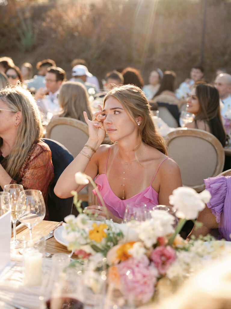 A woman in a pink dress sits at an outdoor event table, wiping her eye. The table is adorned with flowers and glasses. Other guests are seated around her.