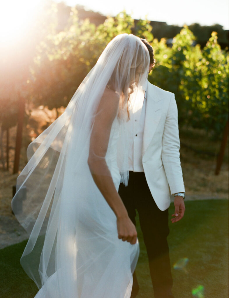 A bride and groom stand outdoors, with the bride in a long veil and white dress, and the groom in a white jacket. They are surrounded by sunlit greenery.