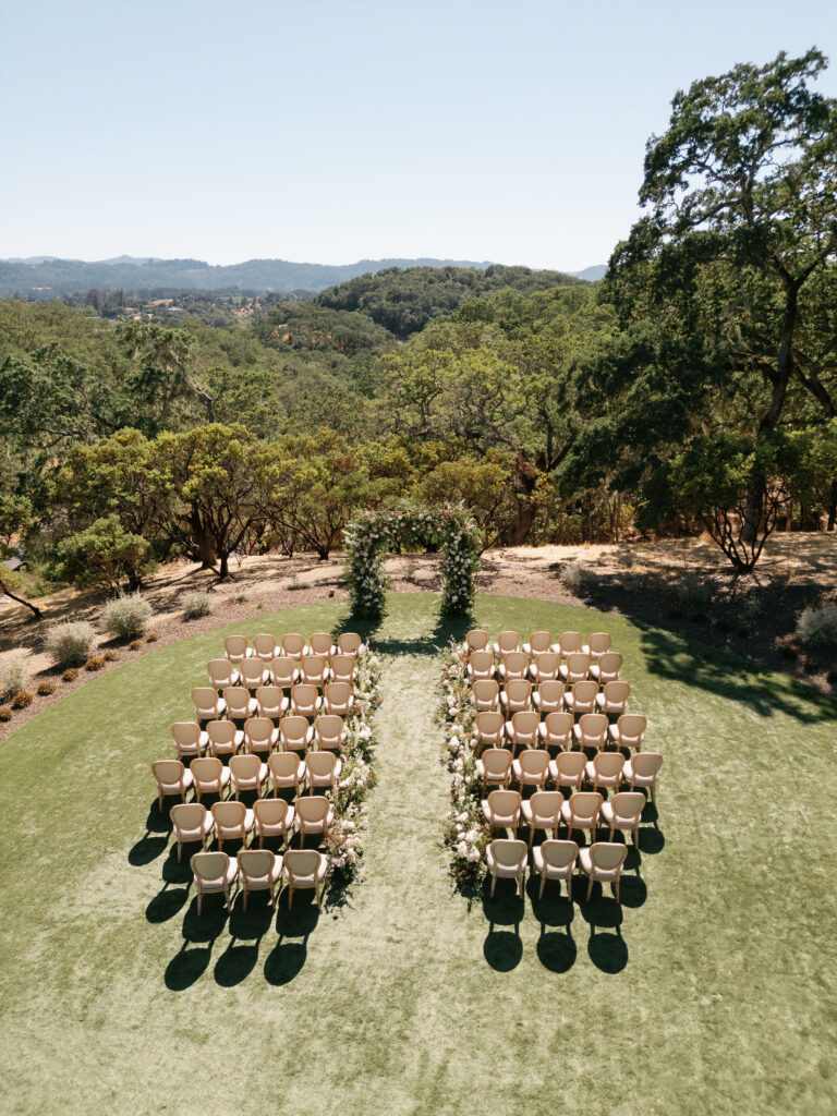Outdoor wedding ceremony setup with rows of chairs on grass facing a floral arch, surrounded by lush greenery and hills in the background.