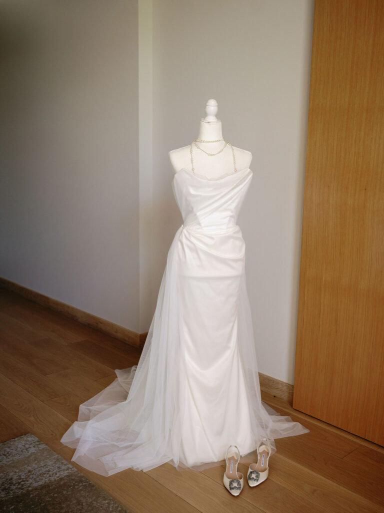 A white strapless wedding dress with a flowing train is displayed on a mannequin. A necklace adorns the neckline, and a pair of silver high-heeled shoes rests on the wooden floor beside it.