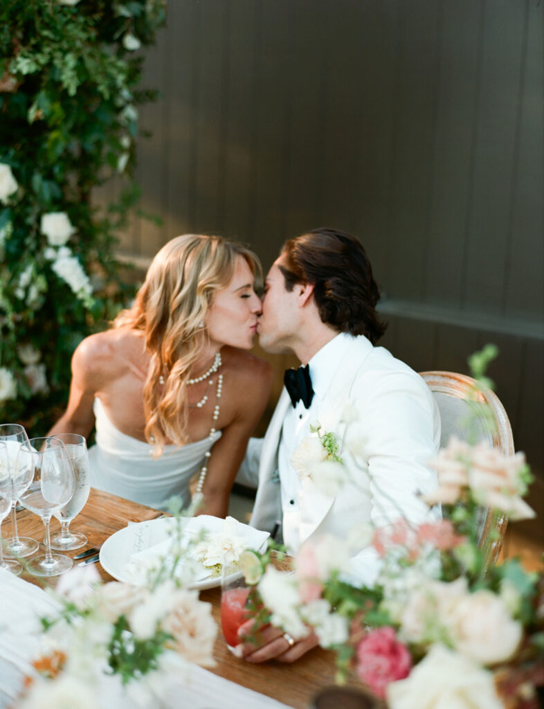 A couple in formal attire kisses at a flower-adorned table set with glassware and plates.