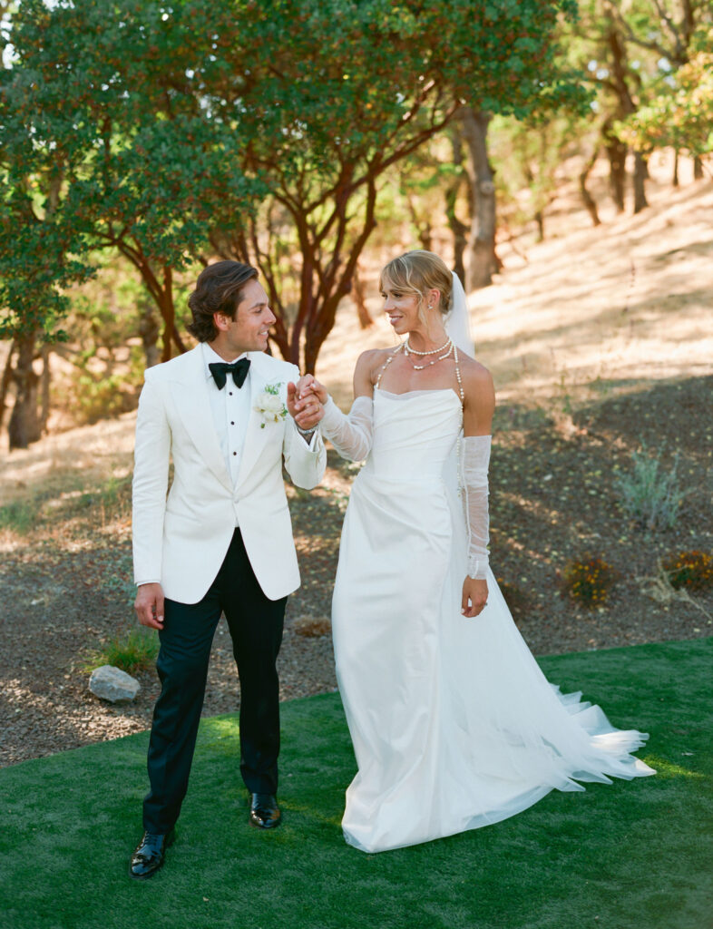 A bride and groom in formal attire walk hand in hand on a grassy area with trees in the background.