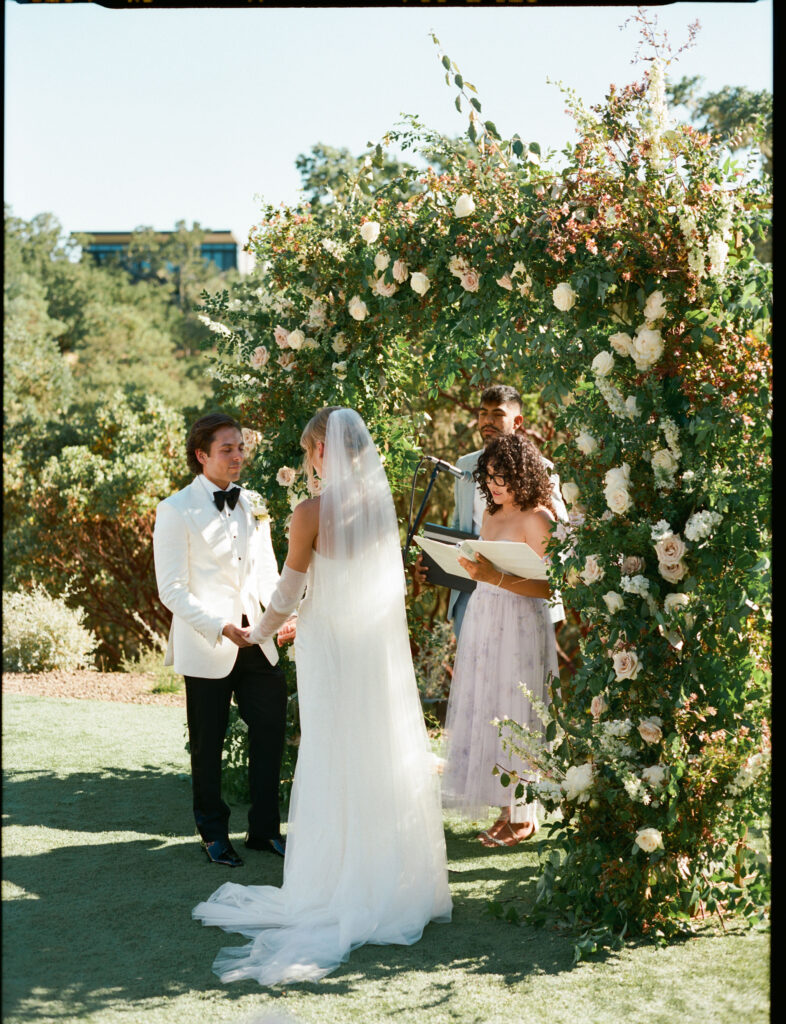 A bride and groom stand under a floral arch during an outdoor wedding ceremony, with an officiant holding a book next to them.