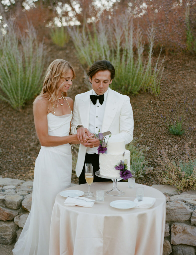 A couple in formal attire cuts a white, two-tiered cake with purple flowers on a small table outdoors.