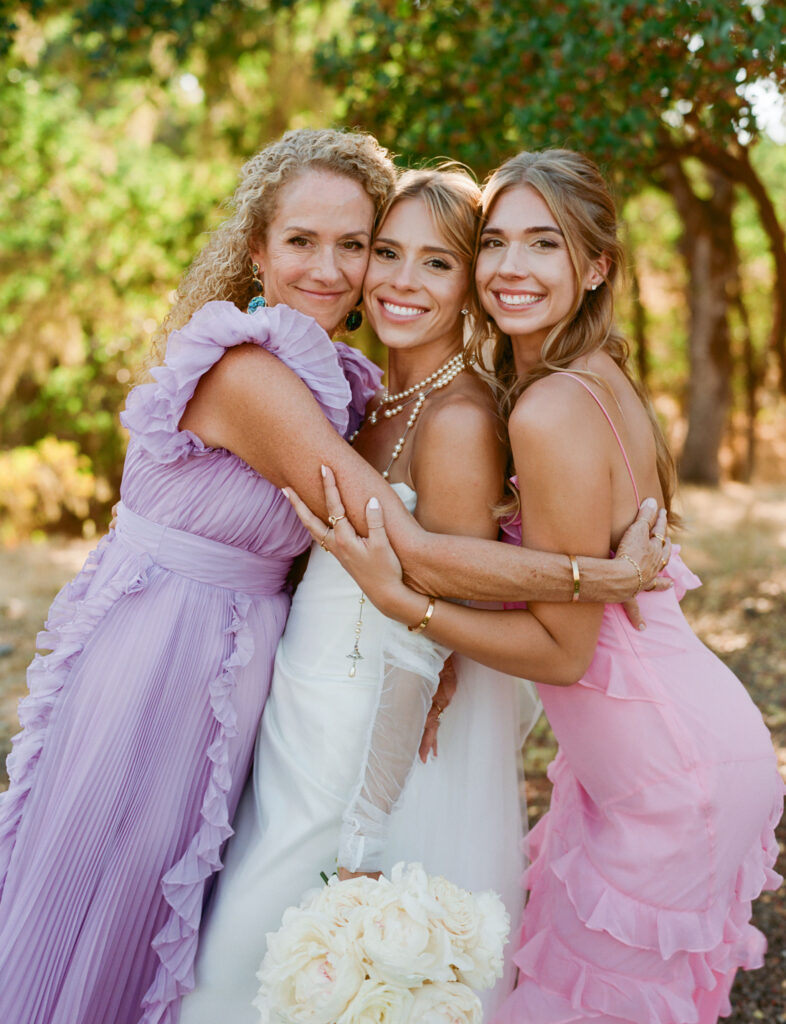Three women in pastel dresses embrace outdoors, smiling at the camera. The woman in the center holds a bouquet of white flowers.