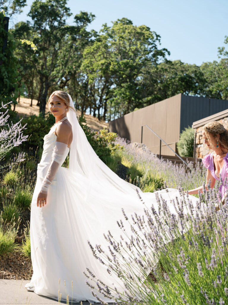 A bride in a white dress and veil stands on a garden path, turning back towards another woman adjusting her train. Lavender plants line the path, with trees and a building in the background.