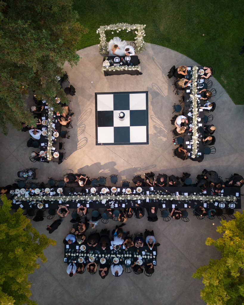Aerial view of a formal outdoor banquet with long tables arranged in an "E" shape, centered around a black and white checkered dance floor, surrounded by greenery.
