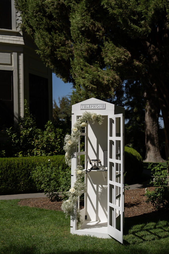 White decorative telephone booth adorned with flowers stands on a grass lawn near trees and a building.