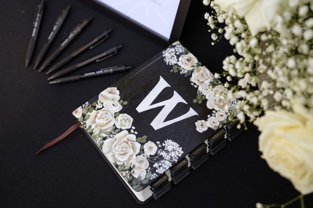 A decorative floral binder with a large letter "W" on the cover sits on a black table next to six black pens and white flowers.
