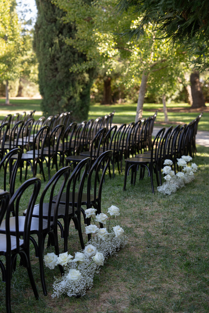 Outdoor wedding setup with rows of black chairs on grass, lined with white flowers. Trees are visible in the background.